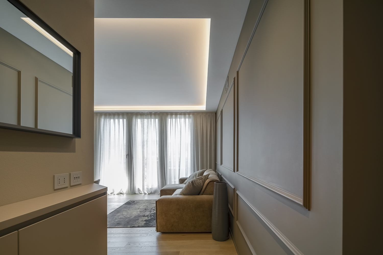 Appartamento C, vista dall’ingresso verso il soggiorno con boiserie chiara, illuminazione indiretta a soffitto e pavimento in legno