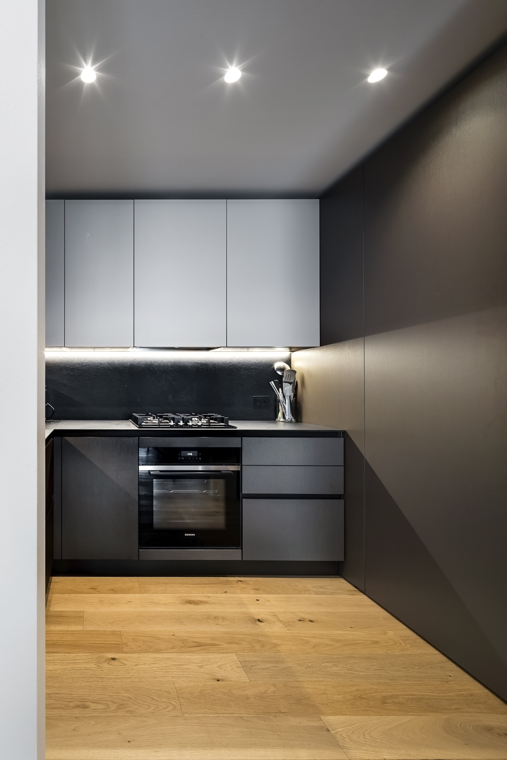 Appartamento C, cucina compatta con finiture scure effetto metallo, pavimento in legno e illuminazione integrata