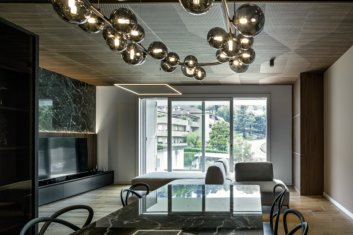 Appartamento YL, zona pranzo e soggiorno con pannelli in noce canaletto a soffitto, illuminazione decorativa e parete in gres effetto marmo Verde Alpi