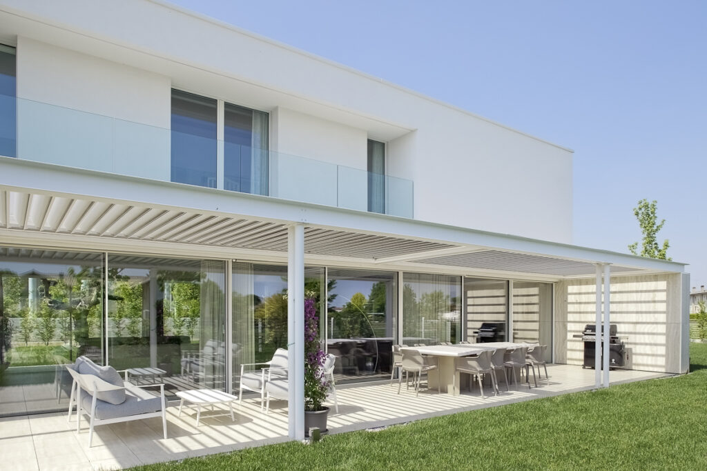 Casa P, portico esterno con pergola bianca, zona pranzo e grandi vetrate verso il giardino